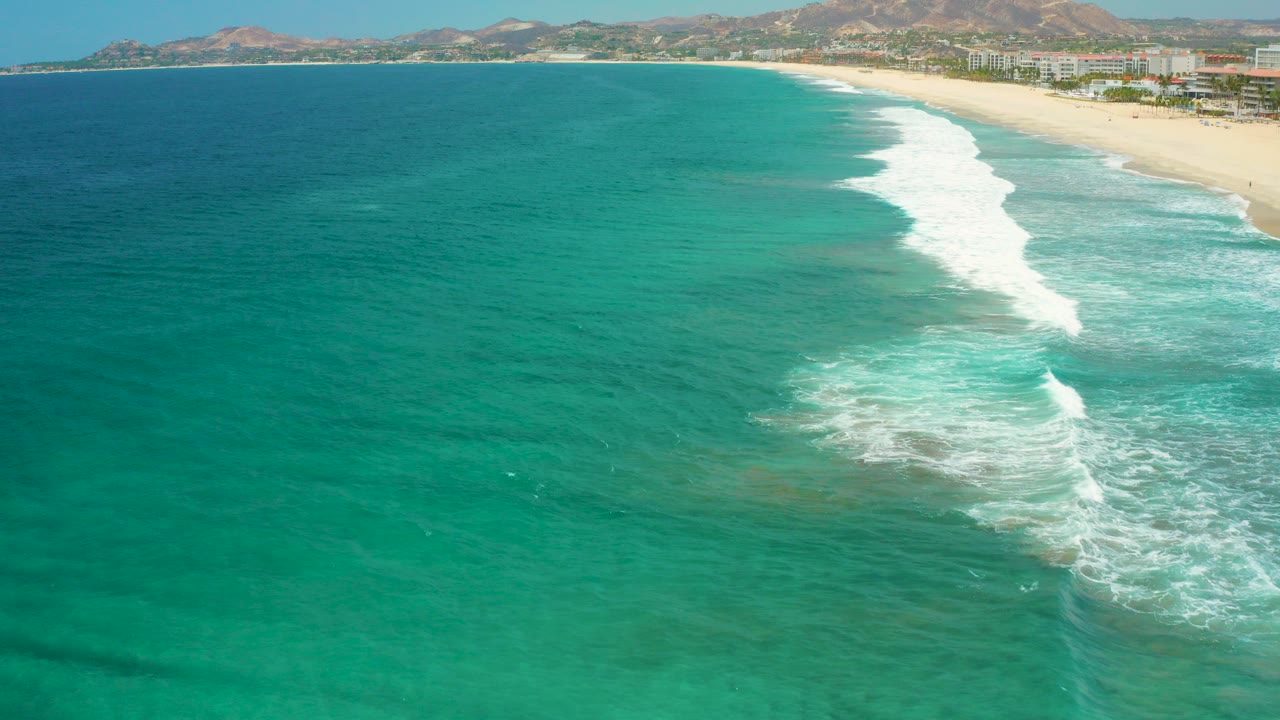 Aerial view over San Jose Del Cabo turquoise water with white surf reaching a sandy beach, shoreline buildings, distant brown hills, and no visible people.
