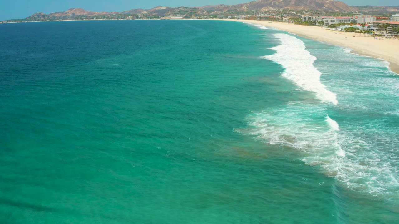 Wide San Jose Del Cabo aerial coastline showing teal water in the foreground, a diagonal white surf line, pale sand, beachfront buildings, hills, and no visible people.