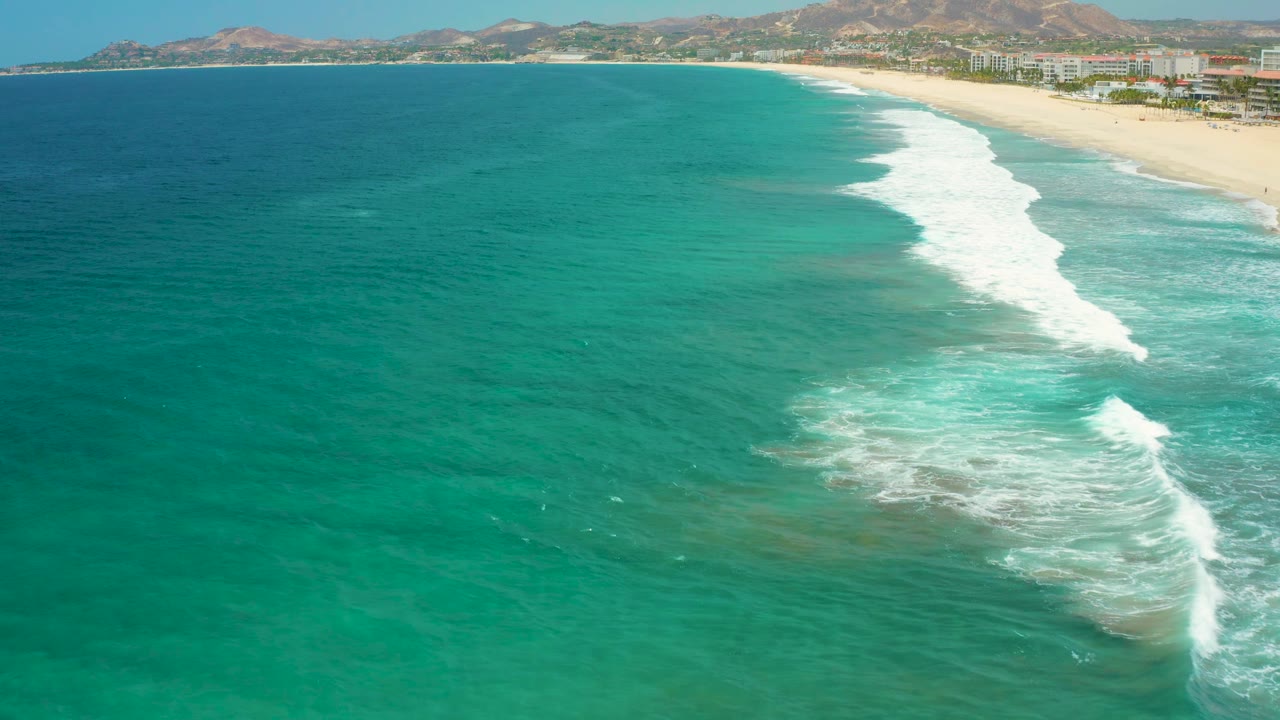 San Jose Del Cabo beach seen from above with a broad teal ocean foreground, white waves breaking close to shore, coastal buildings, dry hills, and no visible people.