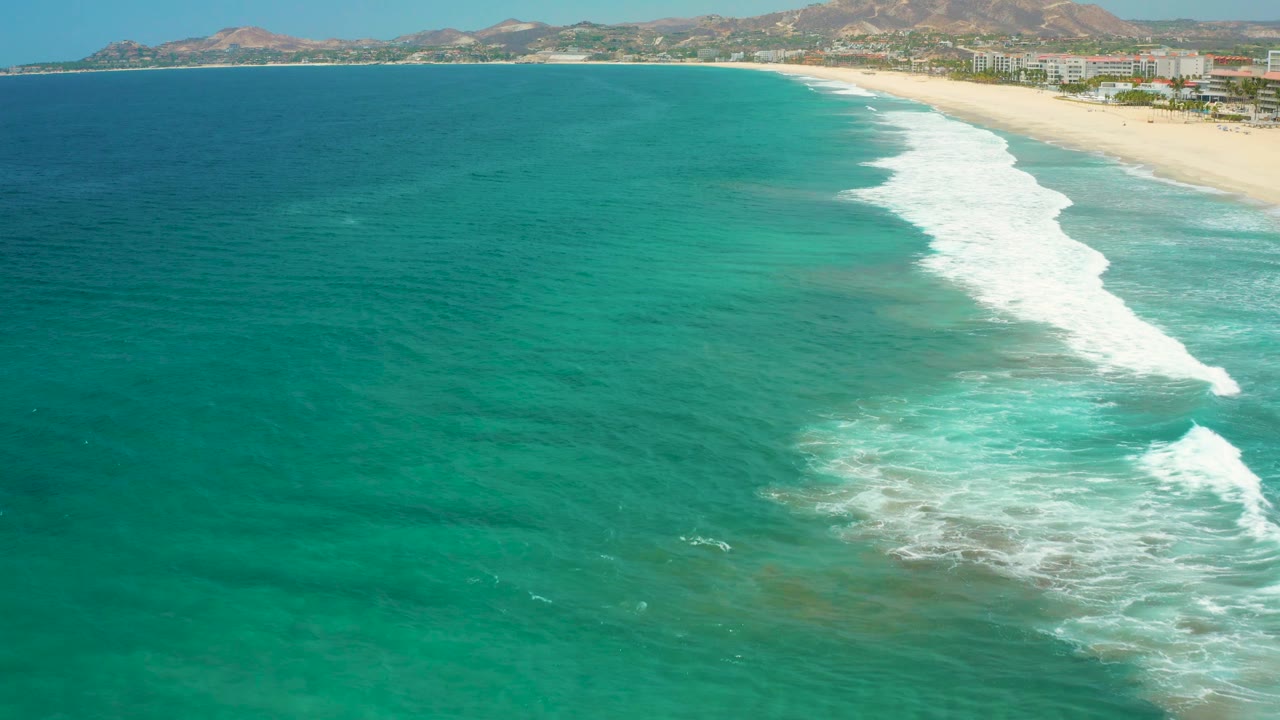 Aerial frame of San Jose Del Cabo shoreline where the beach curves across the top of the image, surf forms a bright band, and no visible people appear.