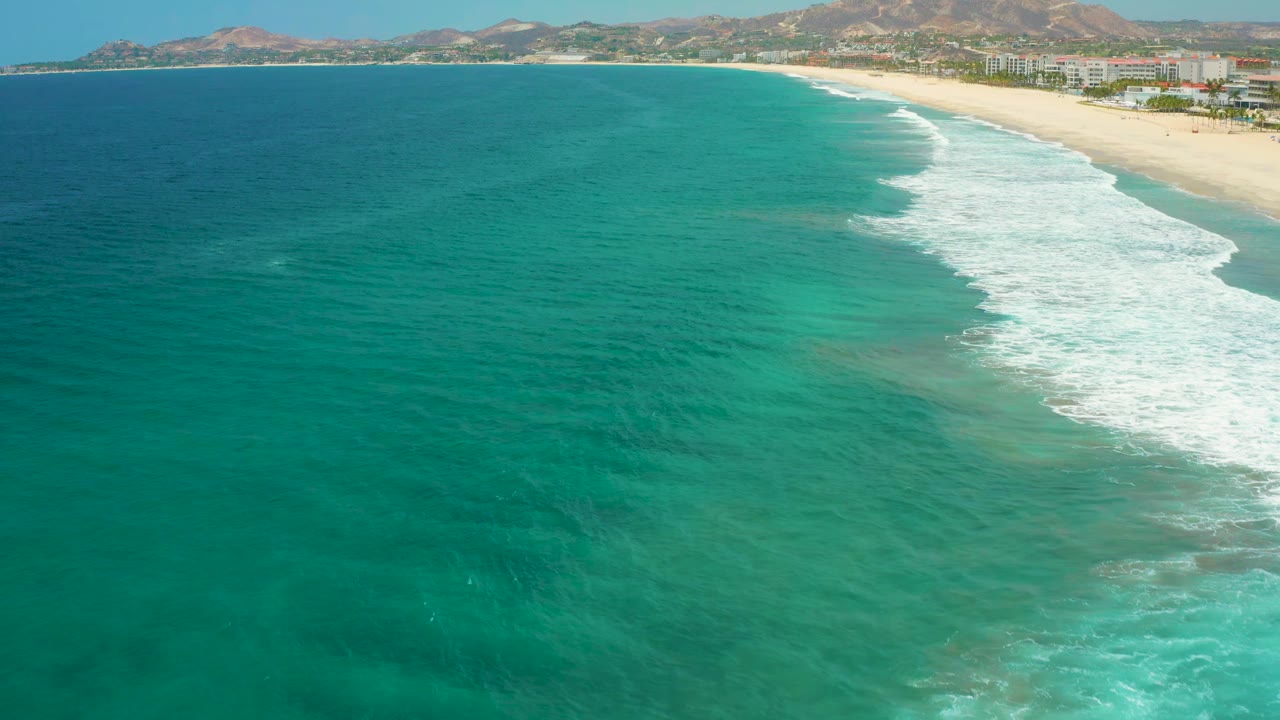 Aerial frame of San Jose Del Cabo, Mexico, showing clear green-blue ocean, a diagonal line of white surf, sandy beach, distant beachfront buildings, mountains, and no visible people.