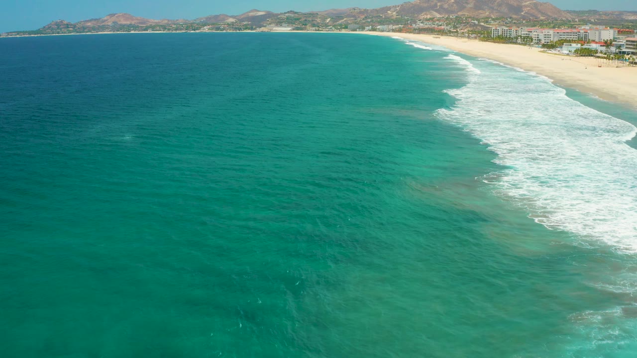 Aerial view in San Jose Del Cabo, Mexico, with broad turquoise water, white surf breaking near the beach, a developed shoreline, low mountains on the horizon, and no visible people.