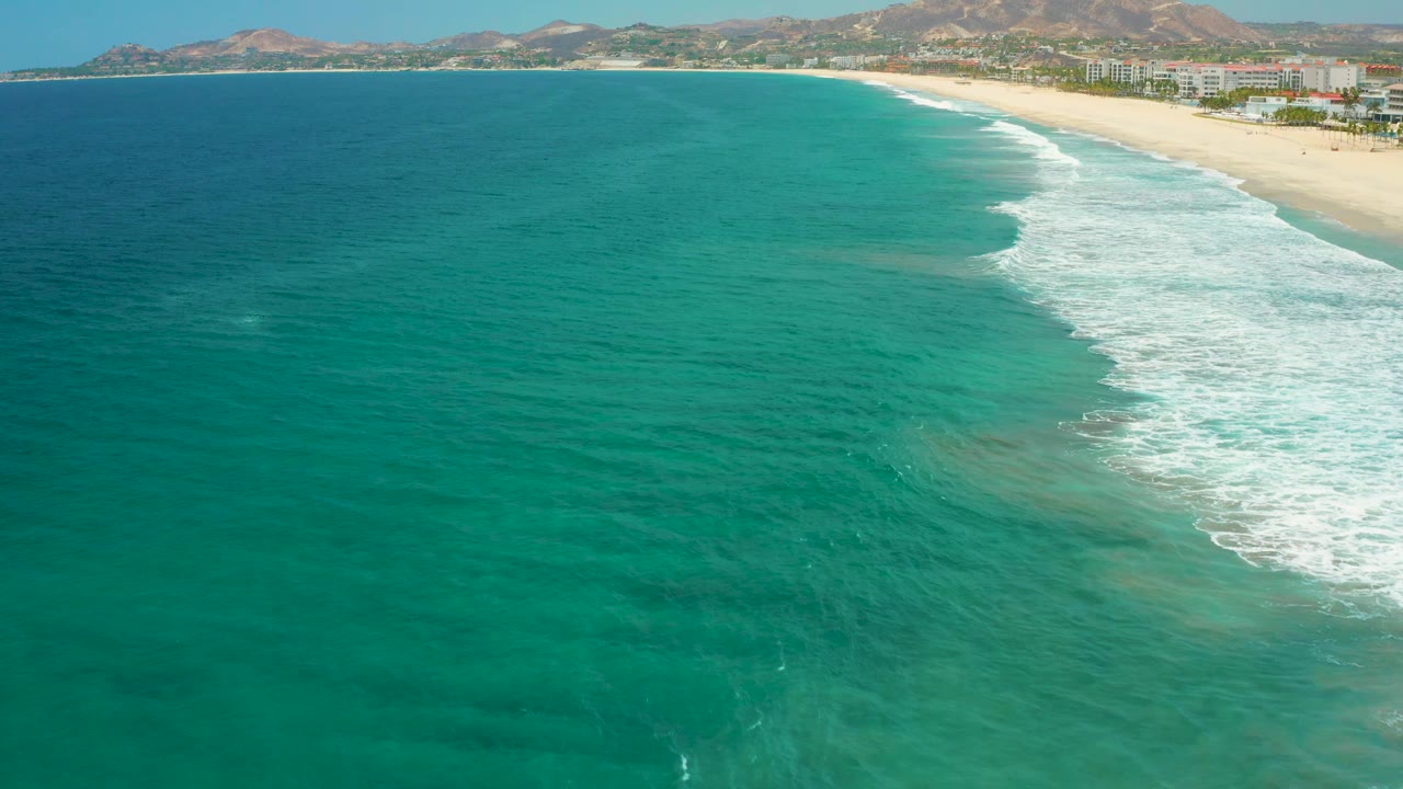 Aerial San Jose Del Cabo, Mexico, frame with teal ocean in front, foamy surf tracing the beach, buildings and trees set back from the sand, dry hills beyond, and no visible people.