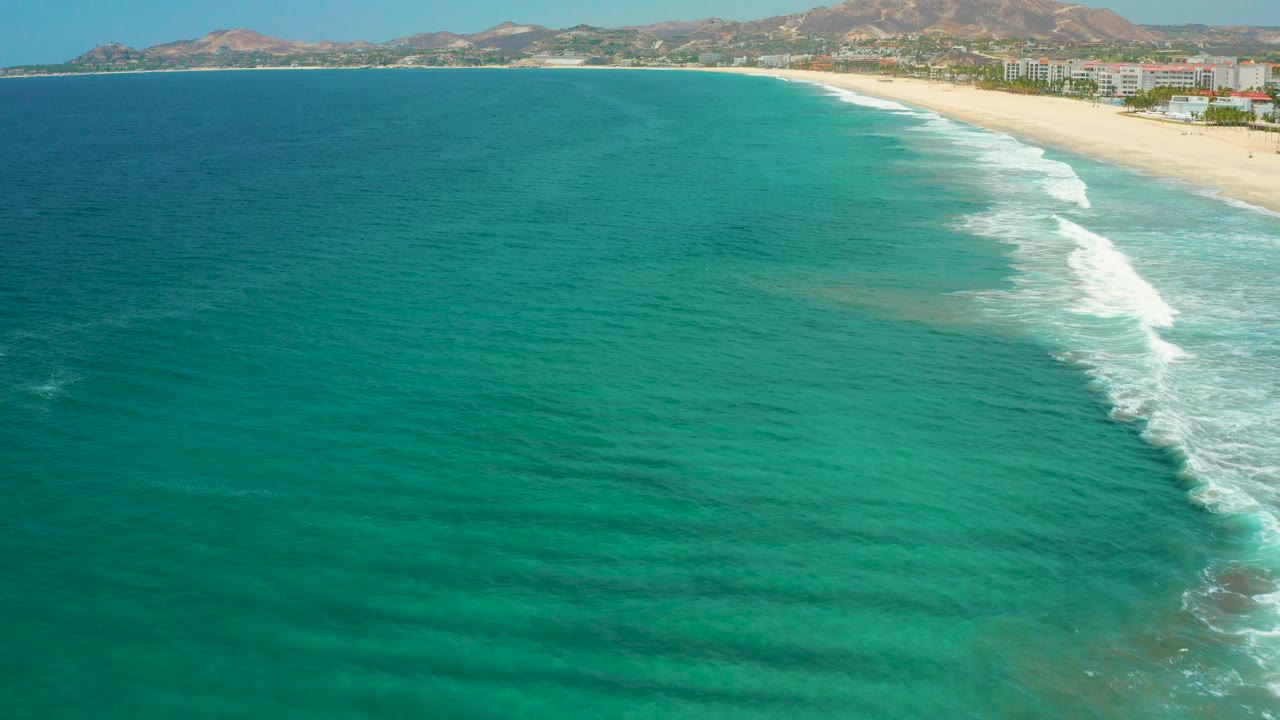Opening aerial frame over turquoise water off San Jose Del Cabo, Mexico, with a pale beach, white surf, shoreline buildings, distant mountains, and no visible people.