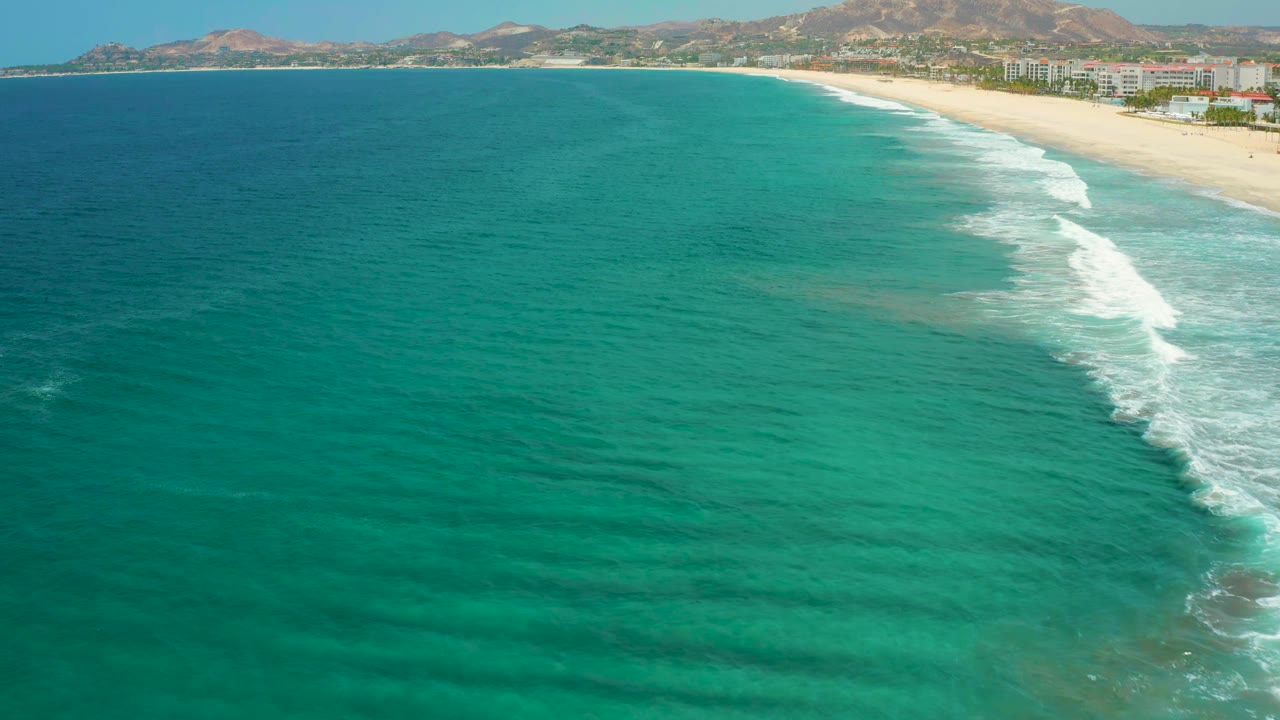 Aerial view one second in, showing clear blue sea stretching across the foreground, a diagonal surf band, San Jose Del Cabo beach, distant buildings, mountains, and no visible people.