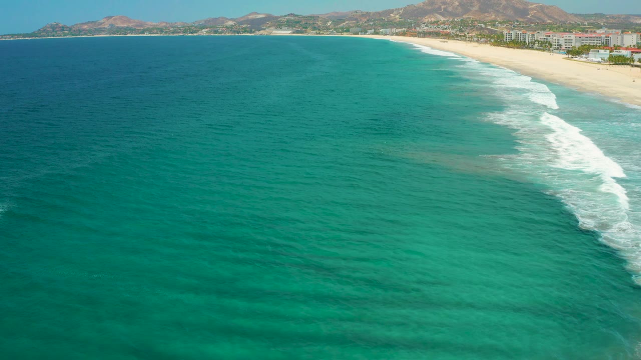 A later aerial frame of San Jose Del Cabo coastline, with more open turquoise water visible, surf breaking near the right edge, a sandy beach beyond it, mountains on the horizon, and no visible people.