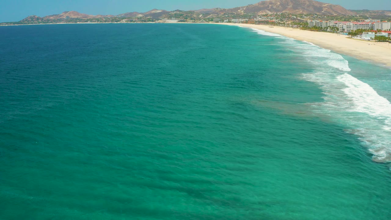 Mid-clip frame over blue-green water near San Jose Del Cabo, Mexico, with white waves approaching the sandy beach, low shoreline development, distant hills, and no visible people.
