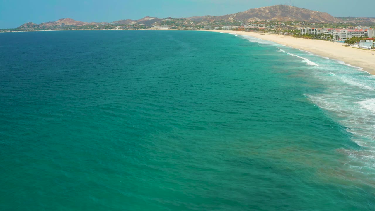 Aerial view over green-blue sea with a diagonal white surf line reaching the San Jose del Cabo beach and low buildings along the shore, no visible people.