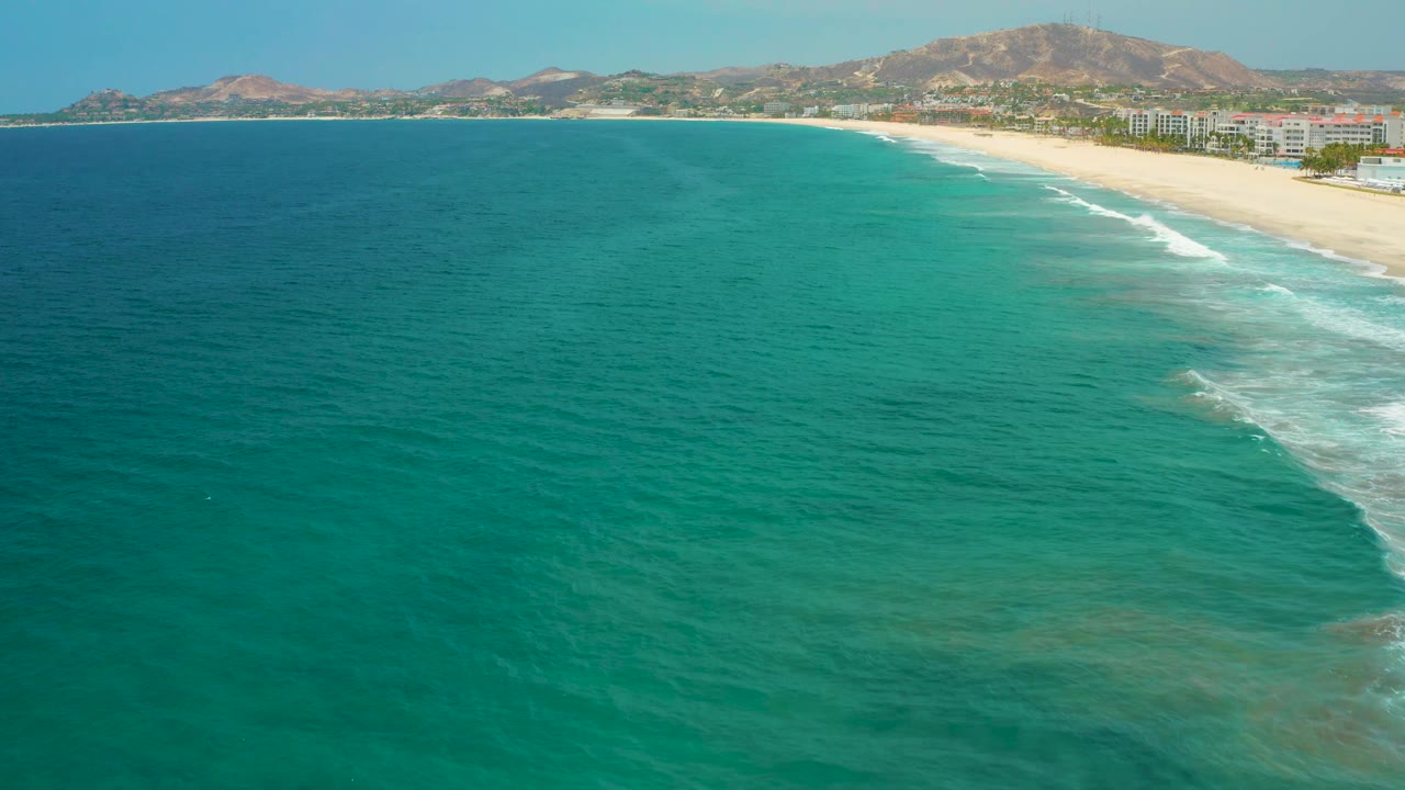 Open turquoise ocean fills the foreground while mild surf breaks along the San Jose del Cabo beach near shoreline buildings and dry hills, no visible people.