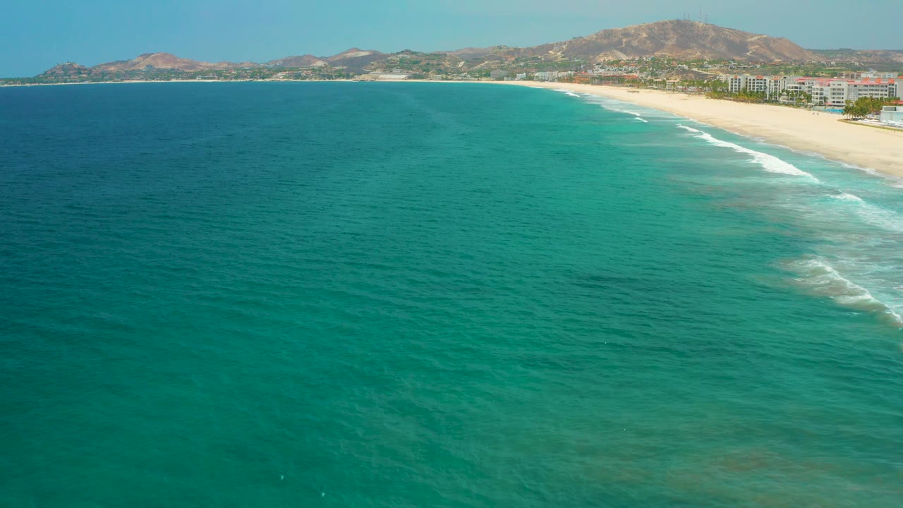 Wide coastal view of calm turquoise water, a curving San Jose del Cabo beach, thin bands of surf, shoreline buildings, and hills under bright sky, no visible people.