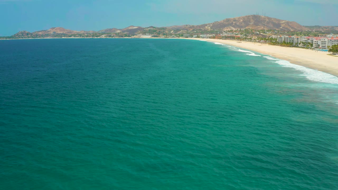 Wide aerial frame of San Jose del Cabo coastline, showing blue-green ocean across the foreground, a pale beach at right, low buildings behind the sand, hills on the horizon, and no visible people.