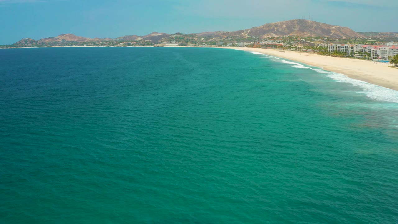High oblique San Jose del Cabo view with textured turquoise water in the foreground, surf tracing the beach at right, a row of distant beachfront buildings, hills beyond, and no visible people.