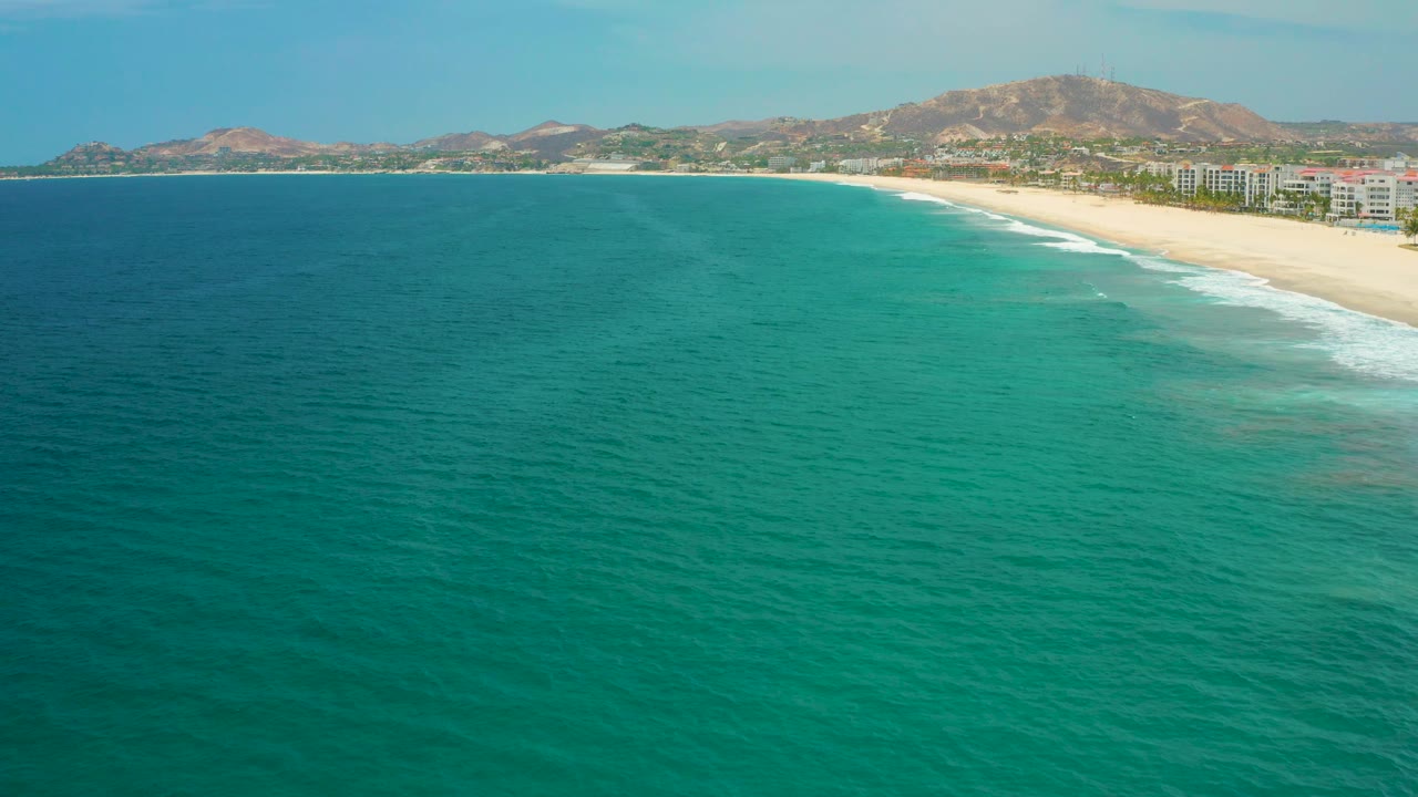 Aerial coastline frame from San Jose del Cabo with open teal ocean across the lower frame, sandy shore to the right, white wave edges, distant buildings, brown hills, and no visible people.