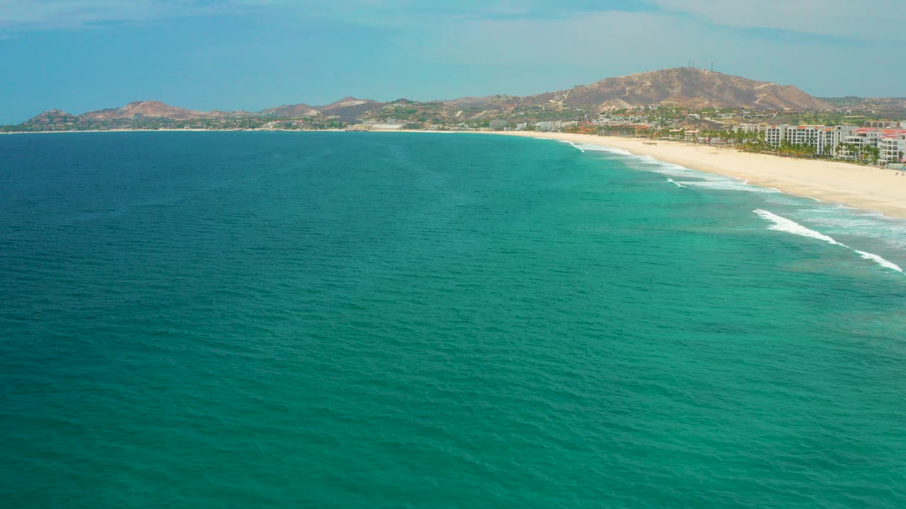 Wide aerial view of blue-green ocean beside a long sandy beach and distant hills.
