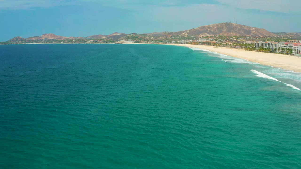 San Jose del Cabo coastline seen from above, with turquoise water filling the lower frame, surf stripes near the sand, shoreline buildings, distant hills, and no visible people.