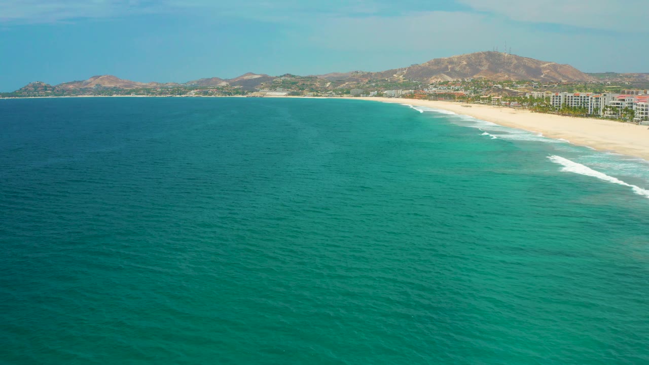 A broad San Jose del Cabo ocean view shows deeper blue water at left, lighter green water near shore, a long beach at right, hillside background, and no visible people.