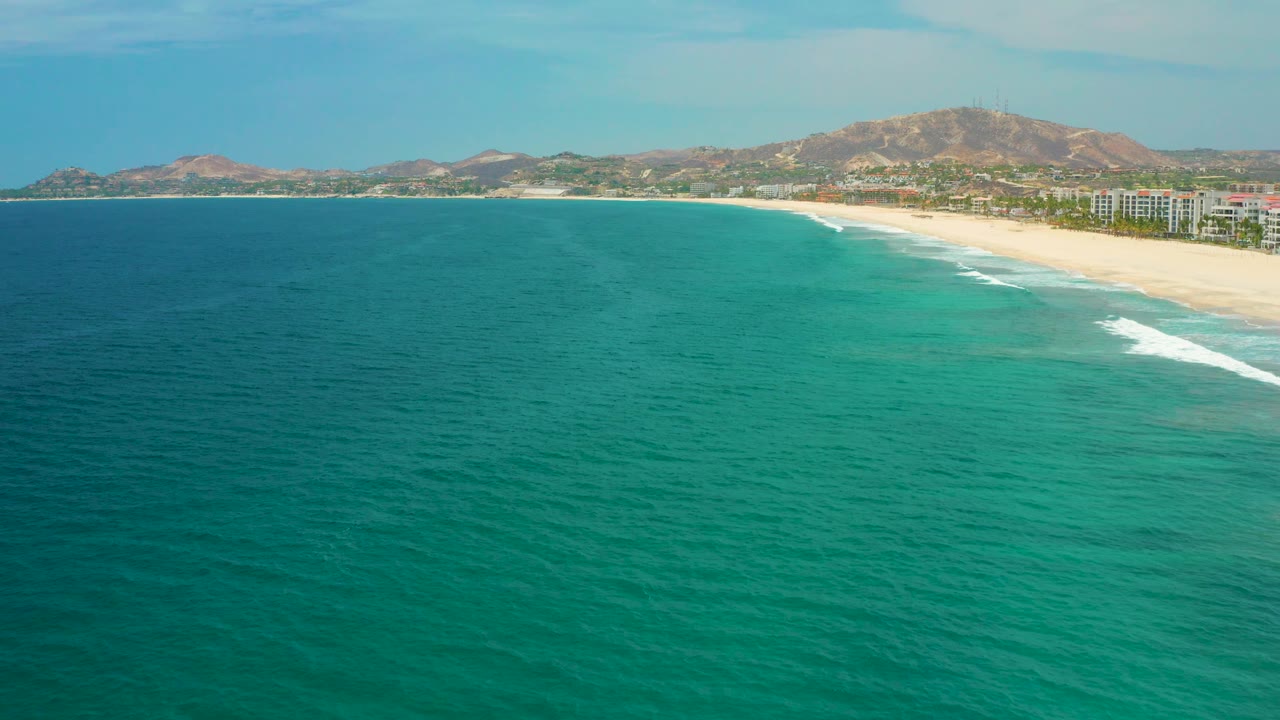 San Jose del Cabo beach appears along the right side as clear blue-green ocean spreads across the foreground, with small white surf, low buildings, dry hills, and no visible people.