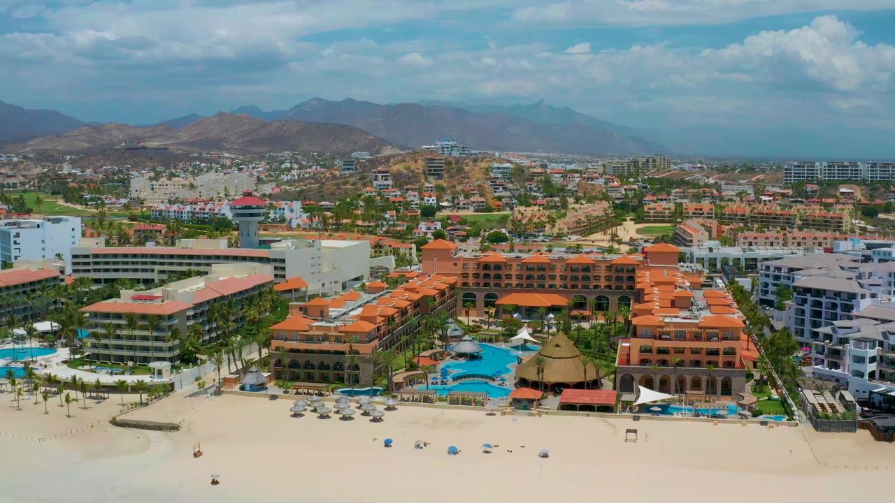 Aerial frame of San Jose Del Cabo showing resort-scale buildings along the sand, turquoise pools, a dense urban midground, mountains, and no visible people.
