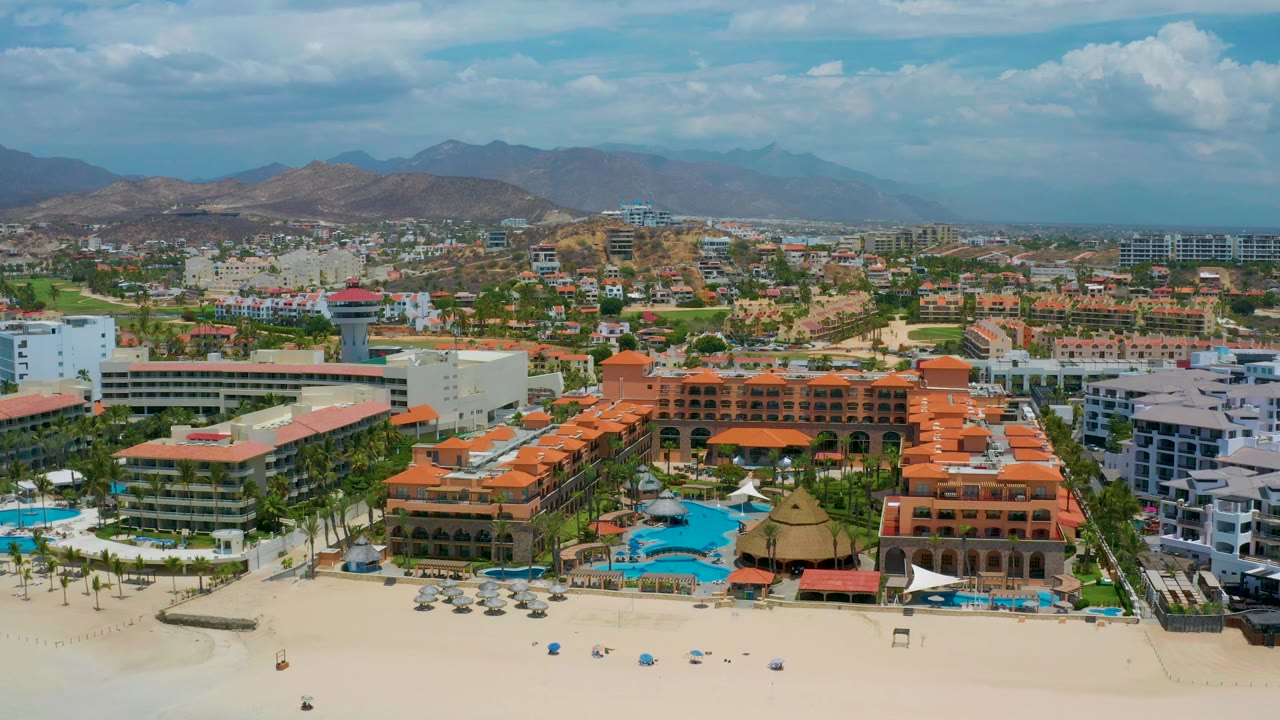 High drone-style view over San Jose Del Cabo with red-roofed and white hotel structures, small pools near the beach, inland buildings, hazy mountains, and no visible people.