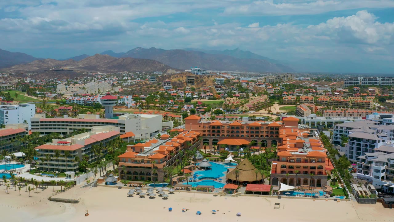 Aerial view across San Jose Del Cabo's beach edge, with a large red-roofed complex, white hotel blocks, blue pool water, inland streets, mountains, and no visible people.