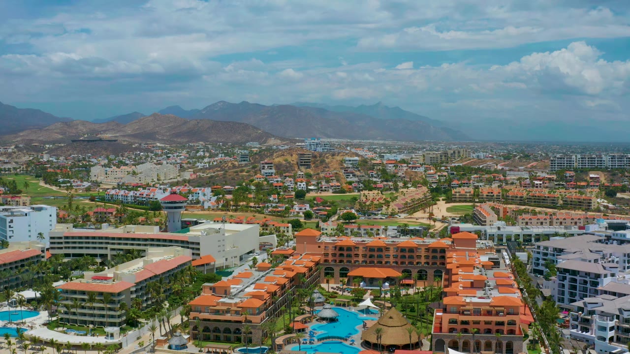San Jose Del Cabo aerial view with white hotel blocks and blue pools in the foreground, green resort grounds and town streets across the middle, hazy mountains in back, and no visible people.