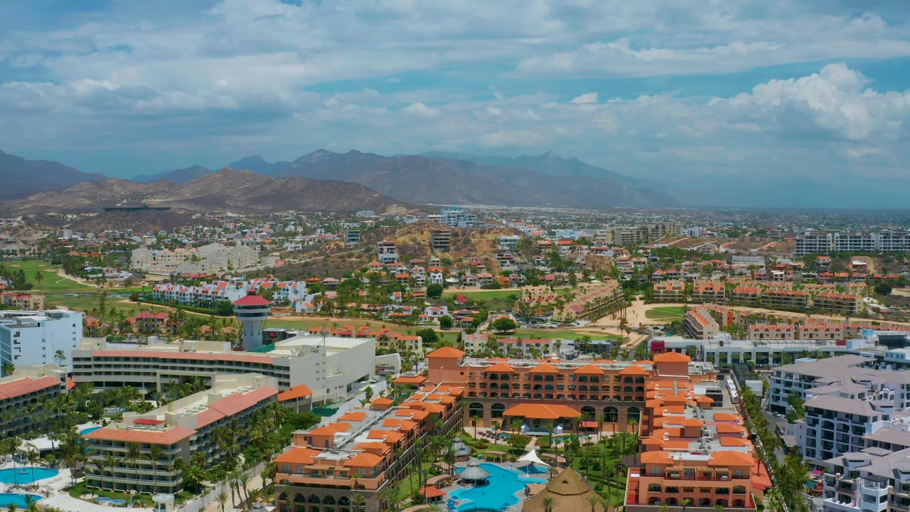 San Jose Del Cabo frame with a white hotel tower and pools in the lower area, orange-roofed resort buildings beside town roads, mountains on the horizon, broad cloud cover, and no visible people.