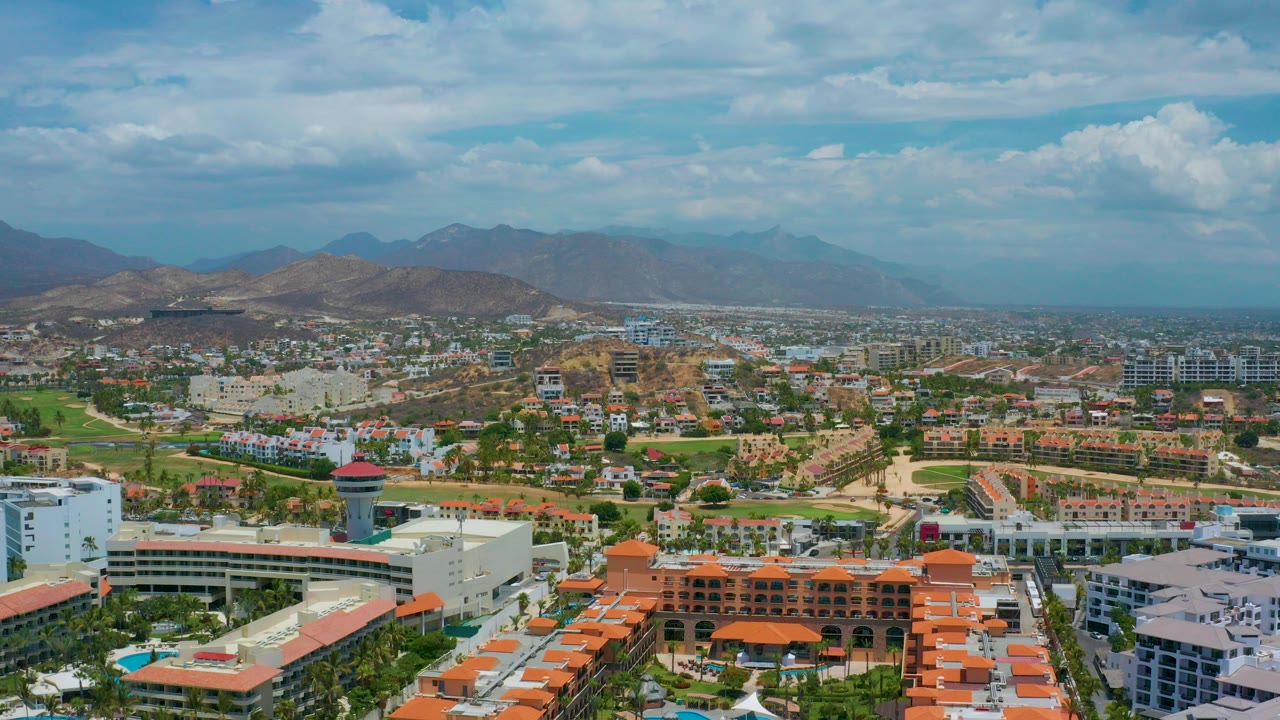 Opening aerial frame over San Jose del Cabo hotel buildings with green grounds, dry hills, hazy mountains, clouded blue sky, and no visible people.