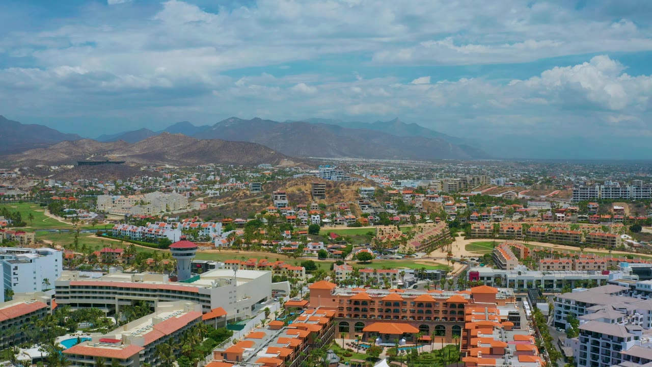 Aerial frame over San Jose del Cabo with white resort blocks and orange rooftops in the foreground, low-rise development and green fields beyond, mountains on the horizon, and no visible people.