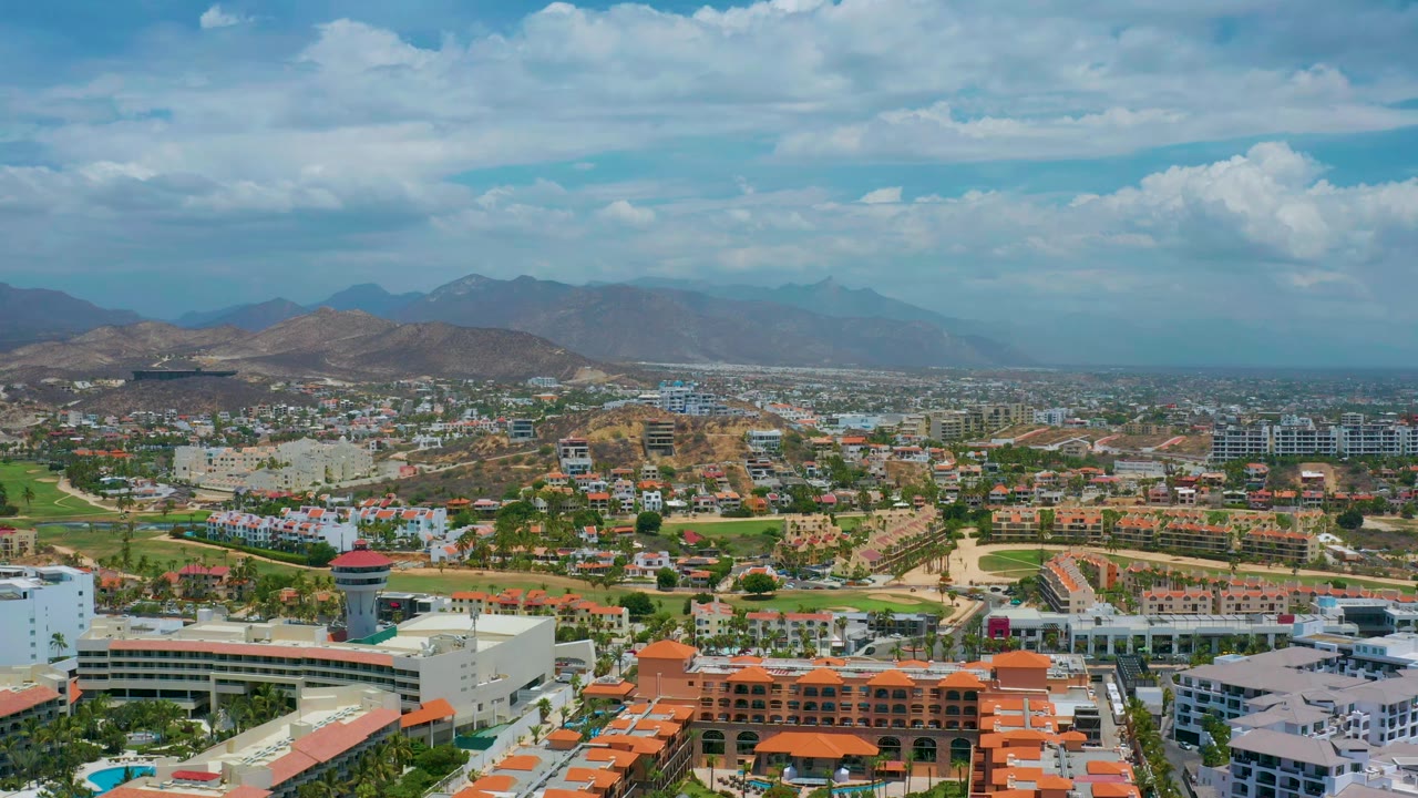 Aerial town view with resort buildings, green spaces, hills, and clouds.