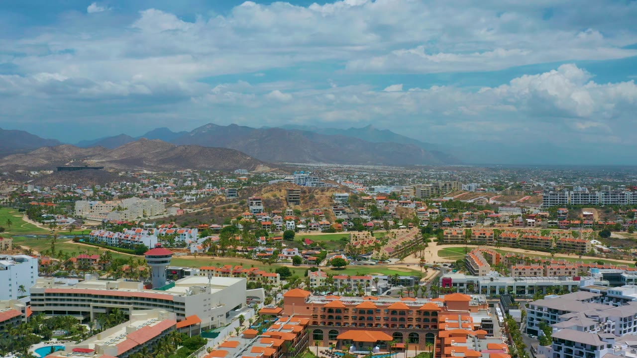 A high aerial frame over San Jose Del Cabo with low-rise development across the foreground, brown hills beyond town, blue sky, scattered clouds, and no visible people.