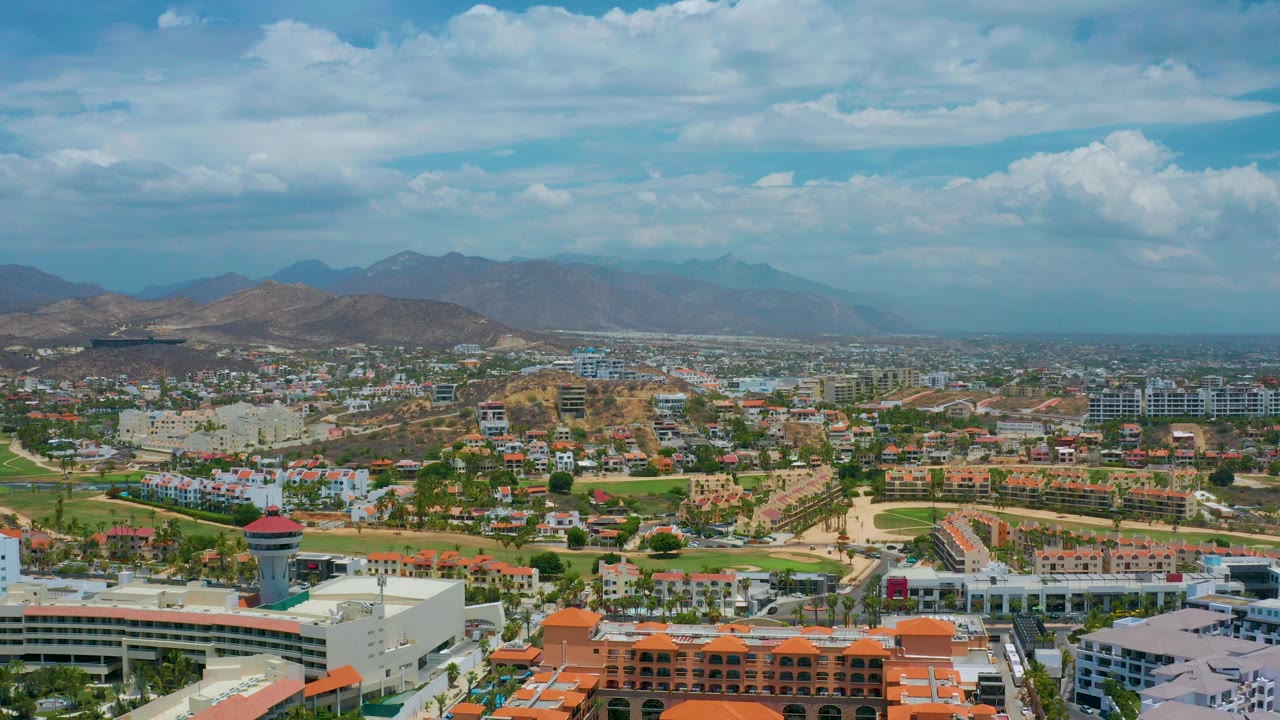 Opening aerial frame over San Jose Del Cabo, Mexico, showing low-rise buildings, hotels, roads, dry hills, hazy mountains, distant sea, and no visible people.