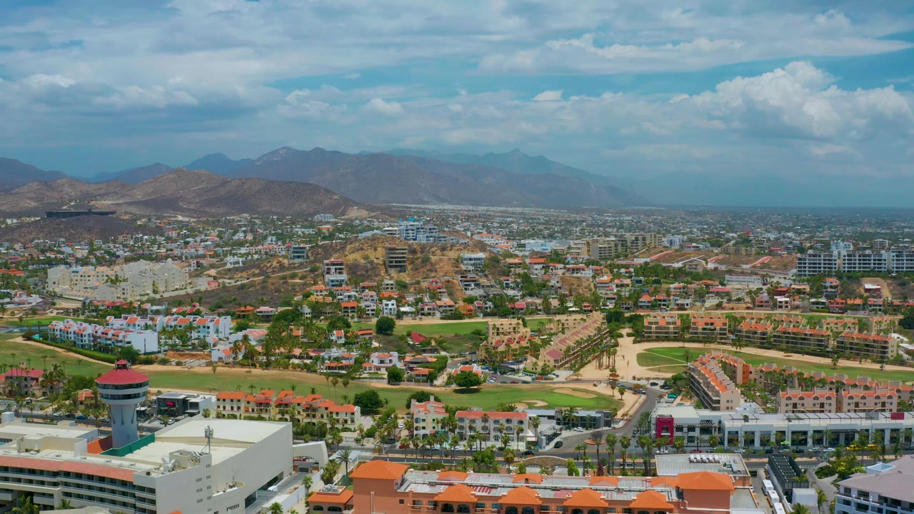 Drone-style wide view across San Jose Del Cabo with a white foreground complex, orange roofs, landscaped green strips, clustered buildings, brown hills, mountains, and no visible people.