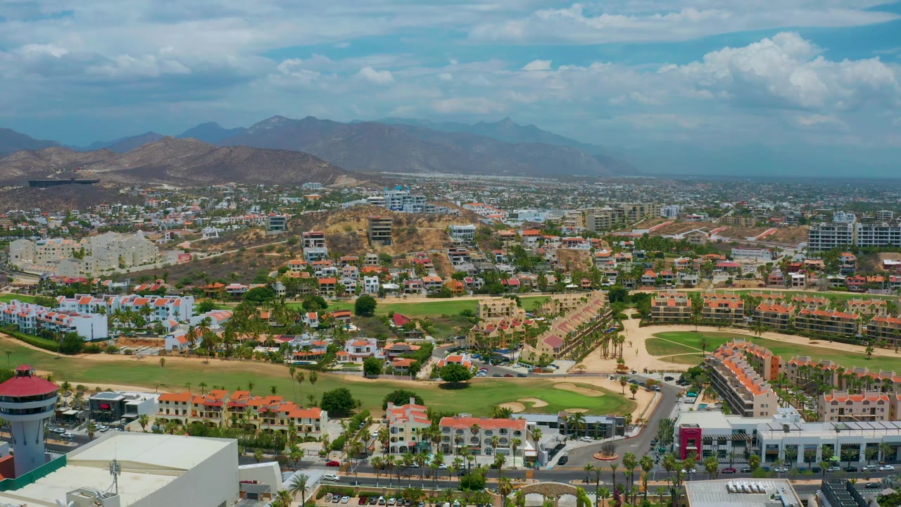 Aerial frame from San Jose del Cabo, Mexico, with white blocks and red roofs in the foreground, green landscaped grounds through the center, dry hills behind town, and no visible people.