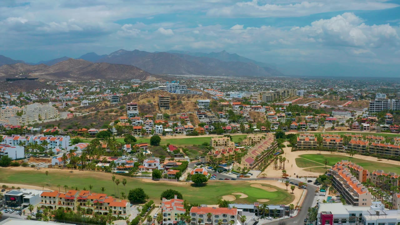 San Jose Del Cabo aerial frame with red-roofed homes, golf-course greens, winding roads, dry hills, distant mountains, blue sky, and no visible people.