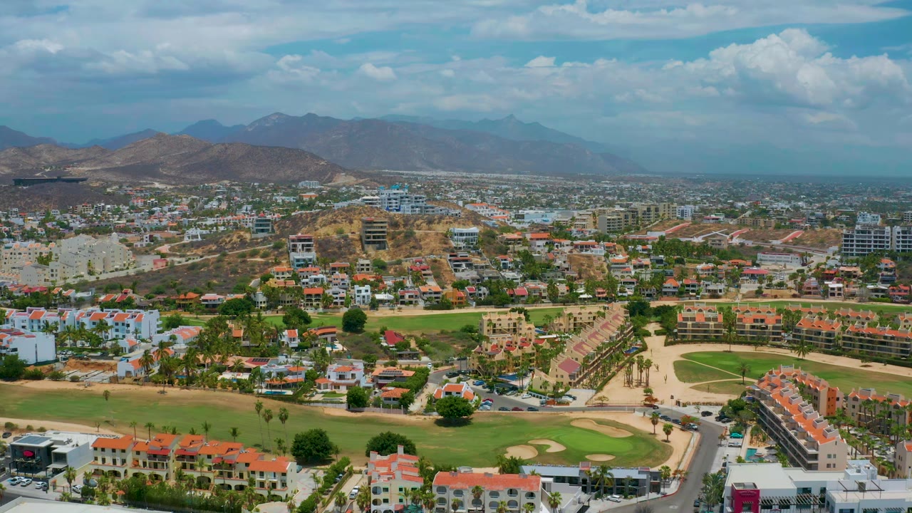 Wide drone view over houses, fairways, roads, hills, and clouded sky