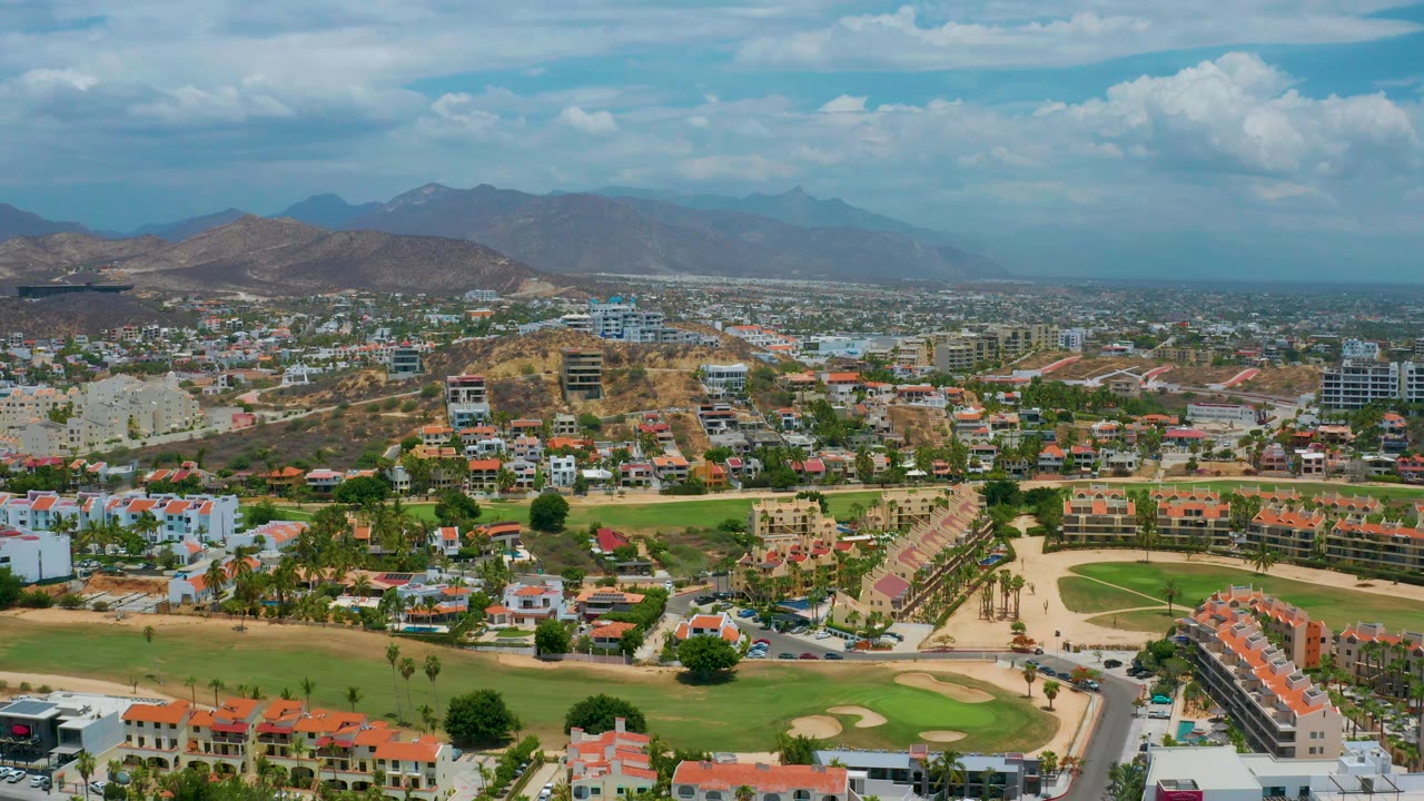 San Jose Del Cabo aerial view showing clustered red roofs, white residential buildings, green lawns, dry surrounding hills, a hazy mountain background, and no visible people.