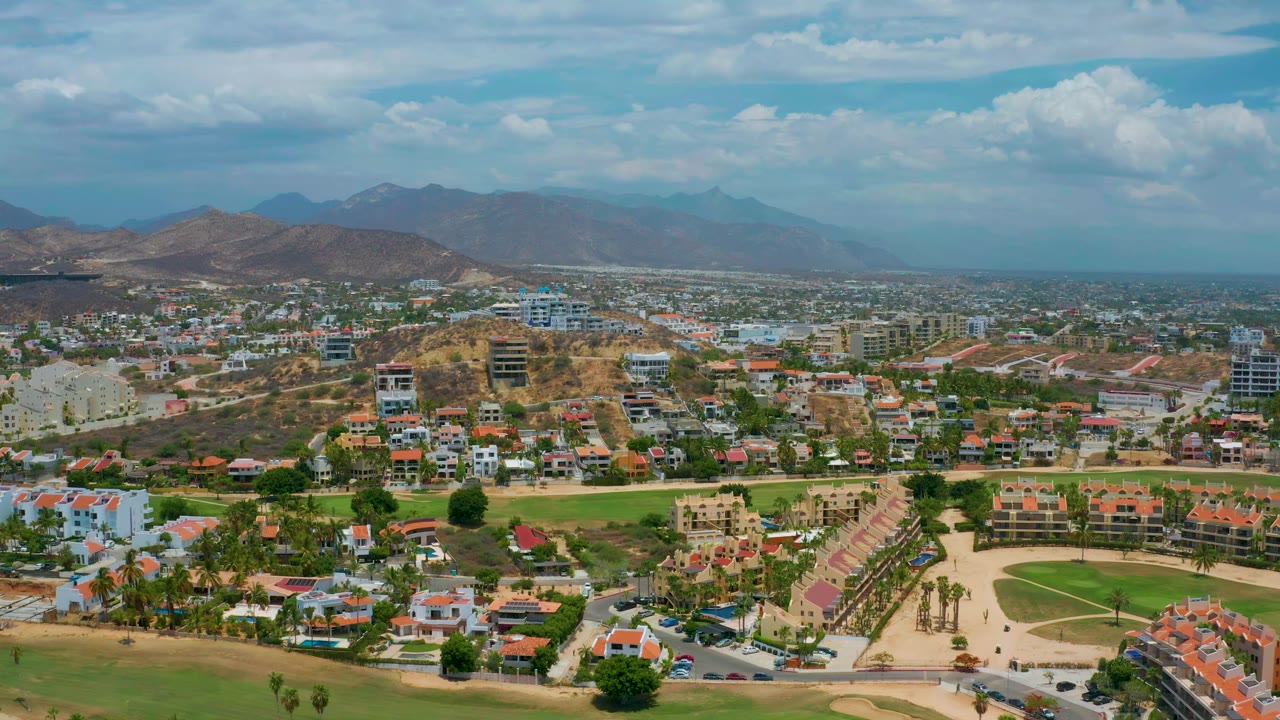 Aerial view over San Jose del Cabo showing compact neighborhoods, resort-style landscaping, tan hills, a hazy mountain ridge, bright sky, and no visible people.