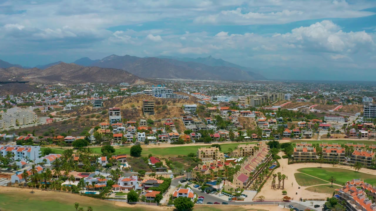 High daytime frame over San Jose del Cabo with a green courtyard area in the lower frame, rows of red roofs, dry hills, distant mountains, and no visible people.