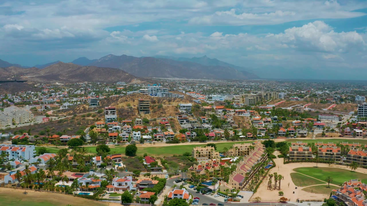 Aerial cityscape with dry hillside, scattered buildings, green lawns, and mountain backdrop.