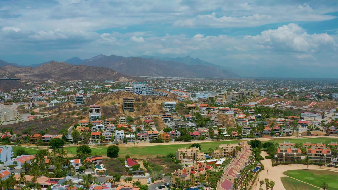 Opening aerial frame of San Jose Del Cabo with residential blocks, a green foreground area, dry hills, distant mountains, clouded blue sky, and no visible people.