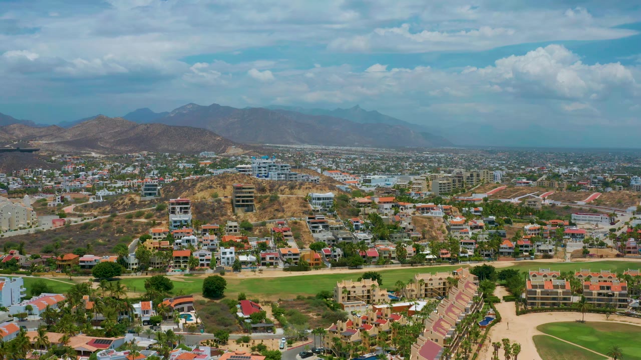 San Jose Del Cabo aerial view one second in, centered on hillside residential buildings, low-rise structures, roads, mountains, and no visible people.