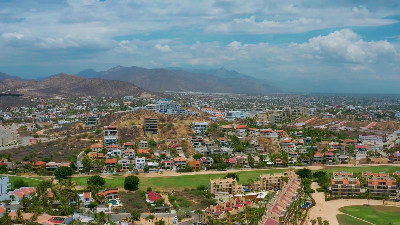 San Jose Del Cabo frame showing rows of homes below tan hills, low-rise urban development, a green foreground strip, distant mountains, and no visible people.