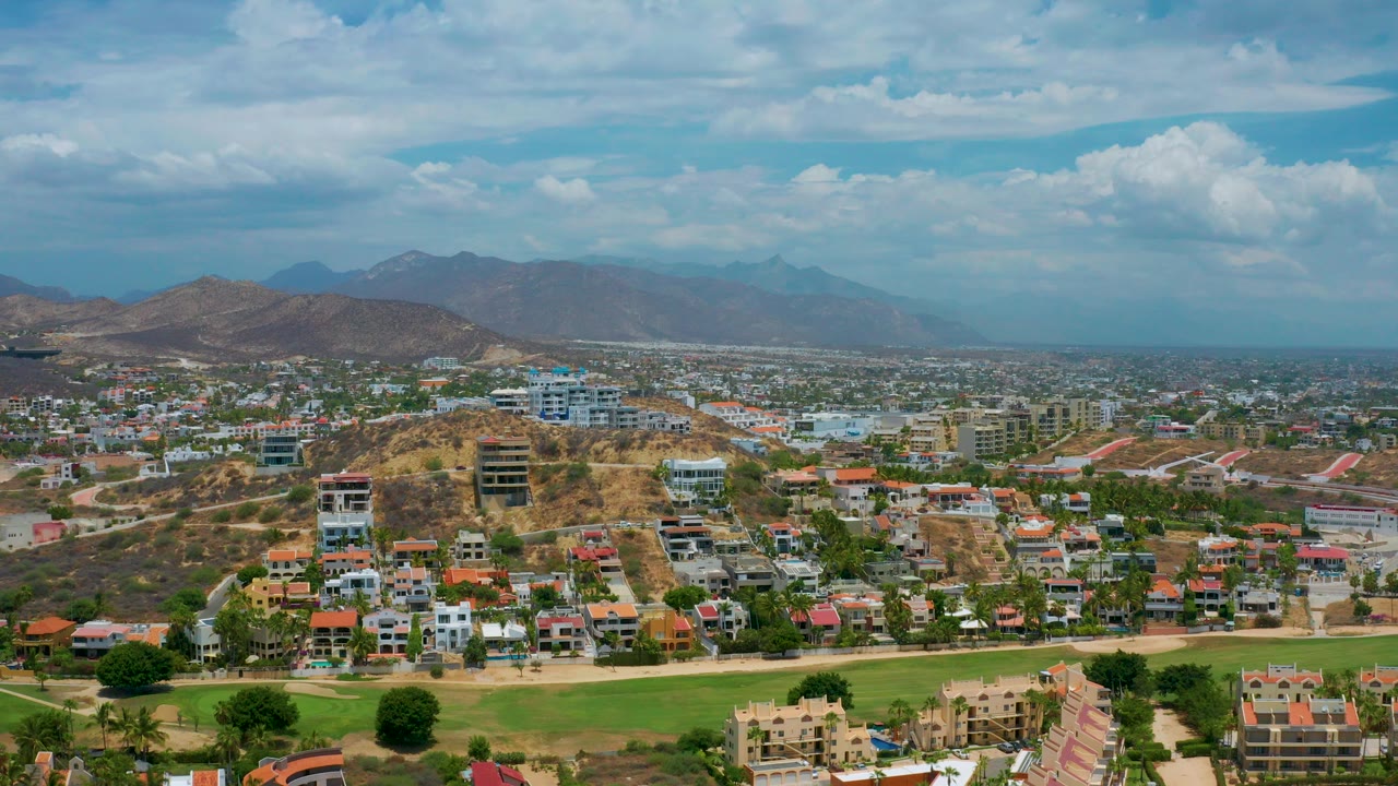 Start of the San Jose Del Cabo aerial view, with low-rise homes, a green foreground strip, dry hillside roads, distant mountains, and no visible people.