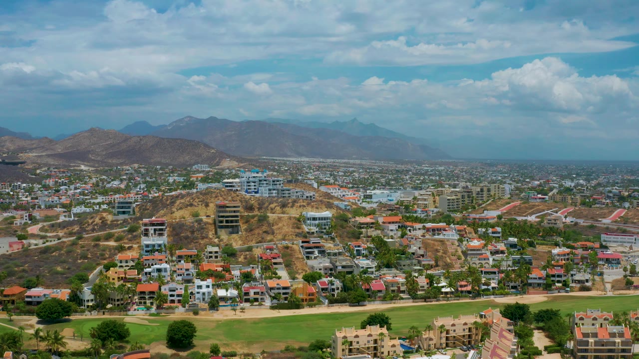 A broad San Jose Del Cabo view shows clustered homes in the foreground and midground, a dry central hill, blue-toned mountains, and no visible people.