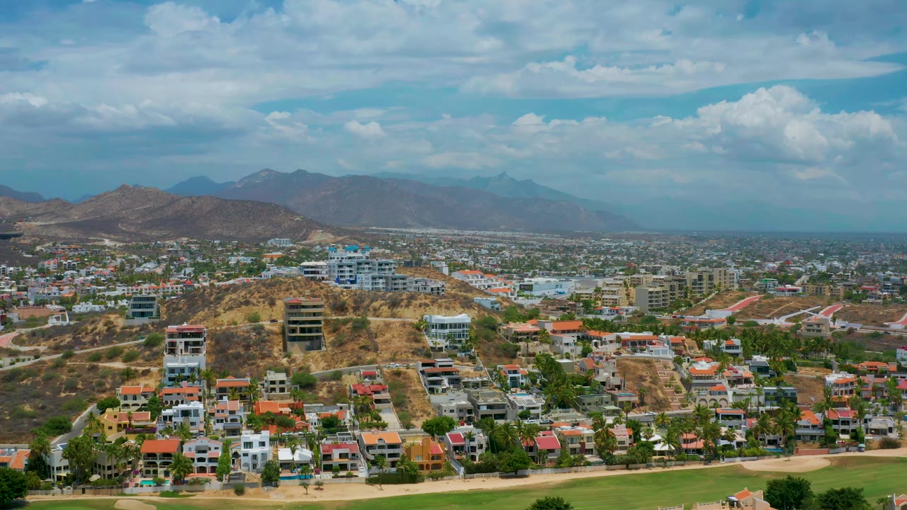 An aerial view of San Jose del Cabo centers on hillside residential buildings, red tiled roofs in the foreground, hazy mountains beyond the town, and no visible people.