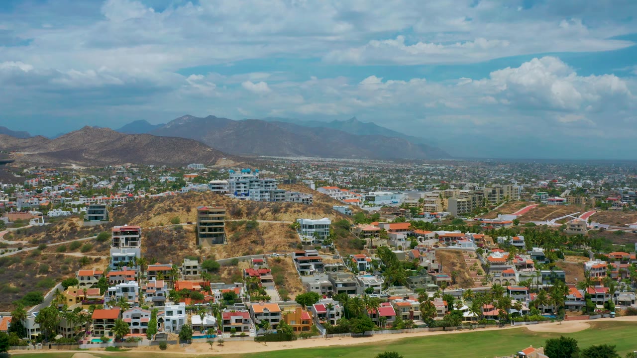 The frame holds a drone-like cityscape of San Jose del Cabo, with a dry hill and modern buildings in the center, rows of homes below, distant mountains, and no visible people.