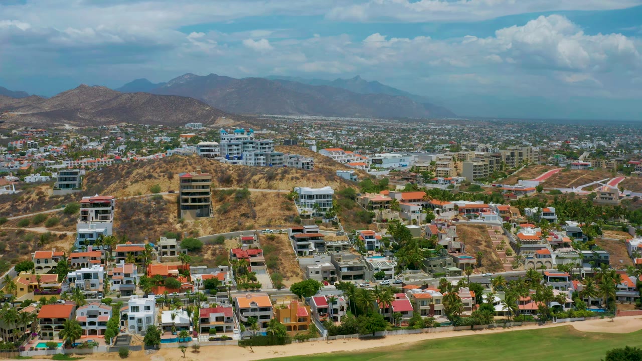 Early aerial view of San Jose Del Cabo showing a dry hillside with modern homes, a broad spread of low buildings, pale roads, mountain ridges on the horizon, and no visible people.