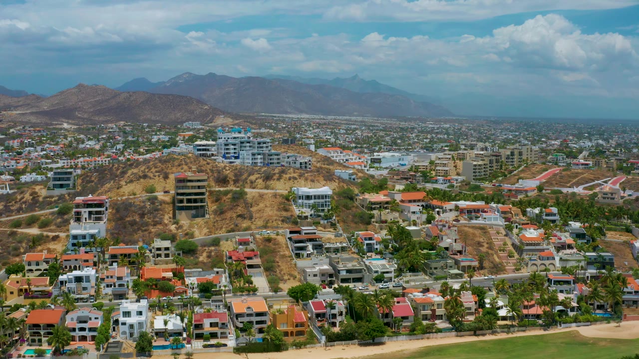Aerial frame over San Jose Del Cabo with red and white rooftops in the lower frame, hillside construction and houses in the center, city sprawl behind them, and no visible people.