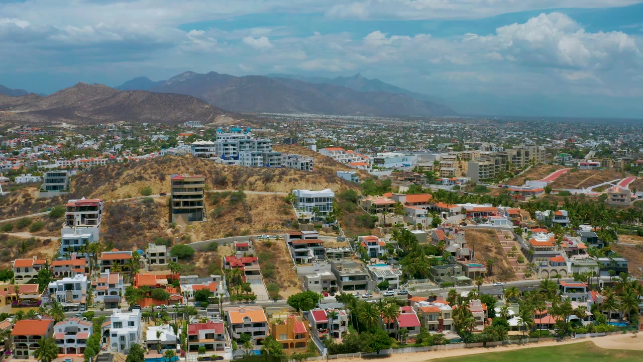 Drone-height view across houses, roads, hills, and distant mountains.