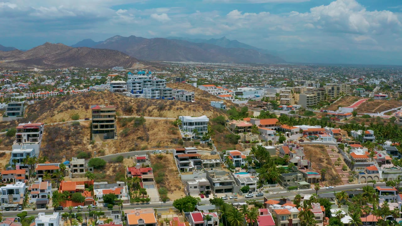 Opening aerial frame of San Jose del Cabo with dry hills, white and tan buildings, red roofs, a broad city grid, distant mountains, and no visible people.
